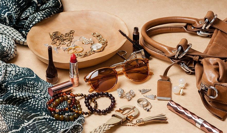 Eine Sonnenbrille, Tasche, Armbänder und weitere Accessoires liegen verteilt auf einem braunen Untergrund